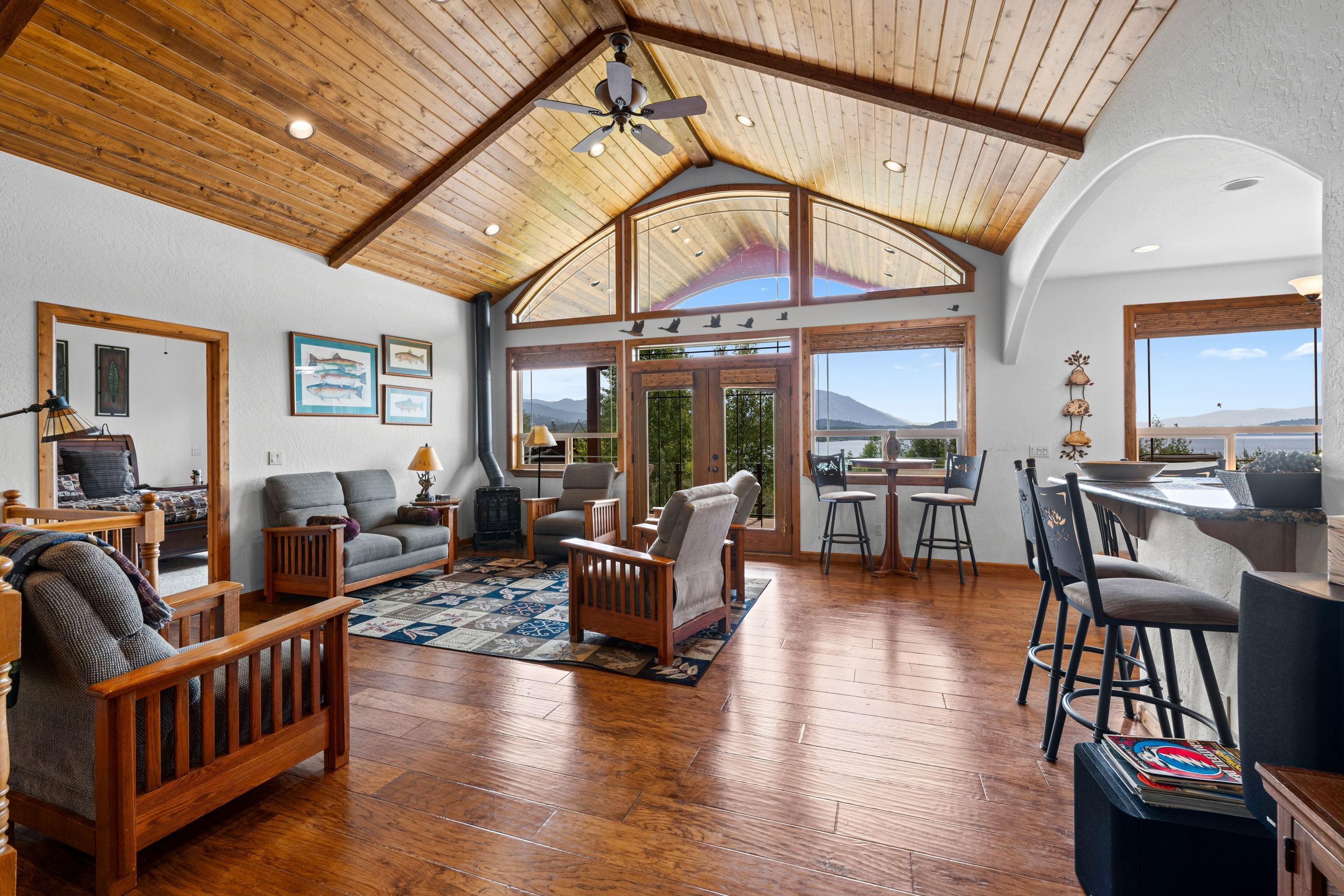 A spacious living area with wooden flooring, a high ceiling, and large windows offering a view of the outdoors.
