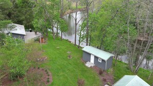 A small, single-story cabin with a metal roof, surrounded by a well-maintained lawn and trees, near a body of water. - The Cottage at River's Bend (Howard)