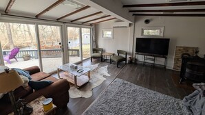A spacious living room with a fireplace, a television, and a large window overlooking a deck. - The Cottage at River's Bend (Howard)