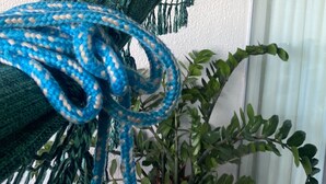 En grön hängmatta är fastknuten vid en vit, strukturerad vägg, en blå lina hänger ner och en krukväxt med gröna blad står i närheten.