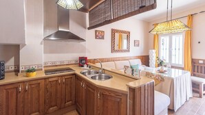 Una cocina con gabinetes de madera, un fregadero de acero inoxidable y una estufa empotrada. Se observa una banca de madera y una mesa con un florero.