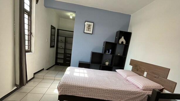 Un dormitorio con cama, escritorio, silla, armario y una pequeña estantería con objetos decorativos.