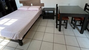 Un dormitorio con una cama, una mesita y una silla. Se observa una estantería a la izquierda.