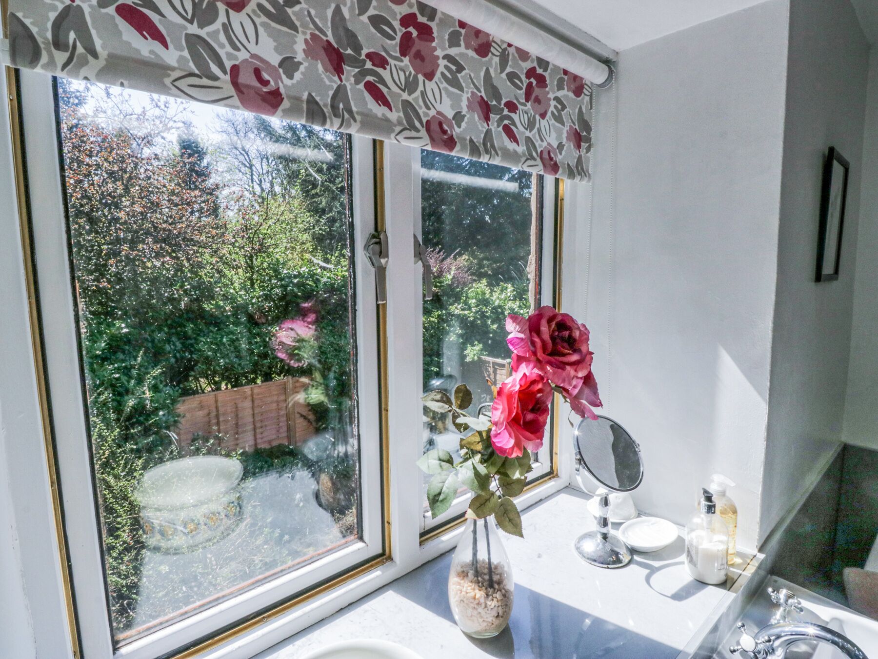 A bathroom with a window, a vase with flowers, a mirror, and toiletries.