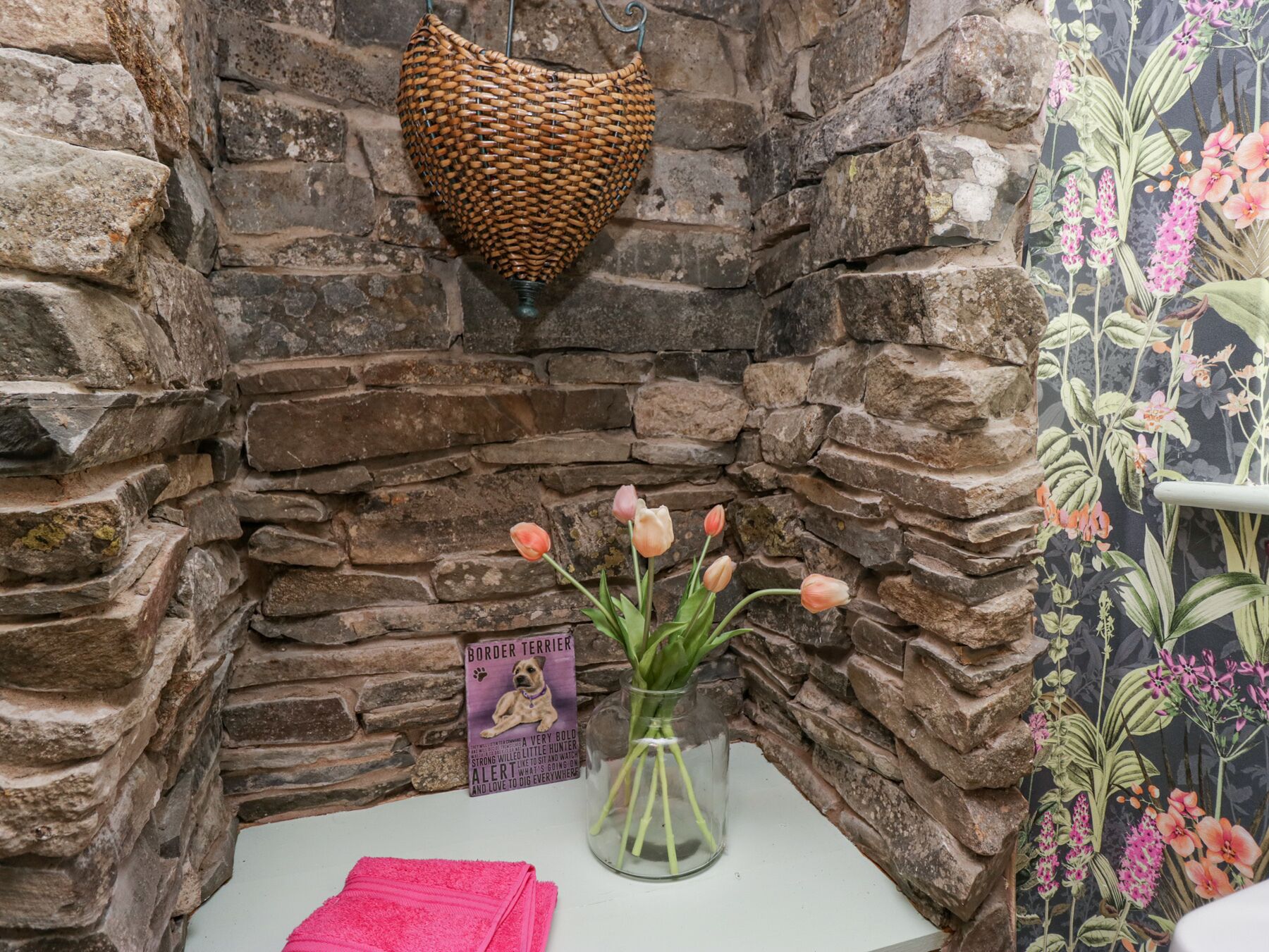 Esquina de pared de piedra con una cesta tejida, un jarrón de cristal con tulipanes, un cartel enmarcado y una toalla rosa.
