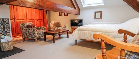 A cozy attic bedroom with a sloped ceiling, a large bed, two patterned armchairs, a wooden bench, a television, and a skylight.