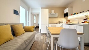 Een moderne woonkamer met een bank, een eettafel en stoelen. Er is een keukenhoek met een magnetron, broodrooster en een mand met fruit.