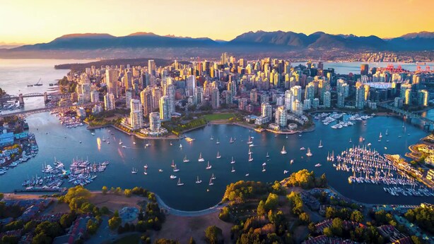 Vista aérea de una ciudad con un puerto deportivo lleno de barcos, rodeado de rascacielos y montañas al fondo.