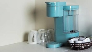 Eine Kaffeemaschine mit blauer Basis und transparentem Wasserbehälter, eine weiße Tasse sowie eine Korb mit Tassen und Deckeln.