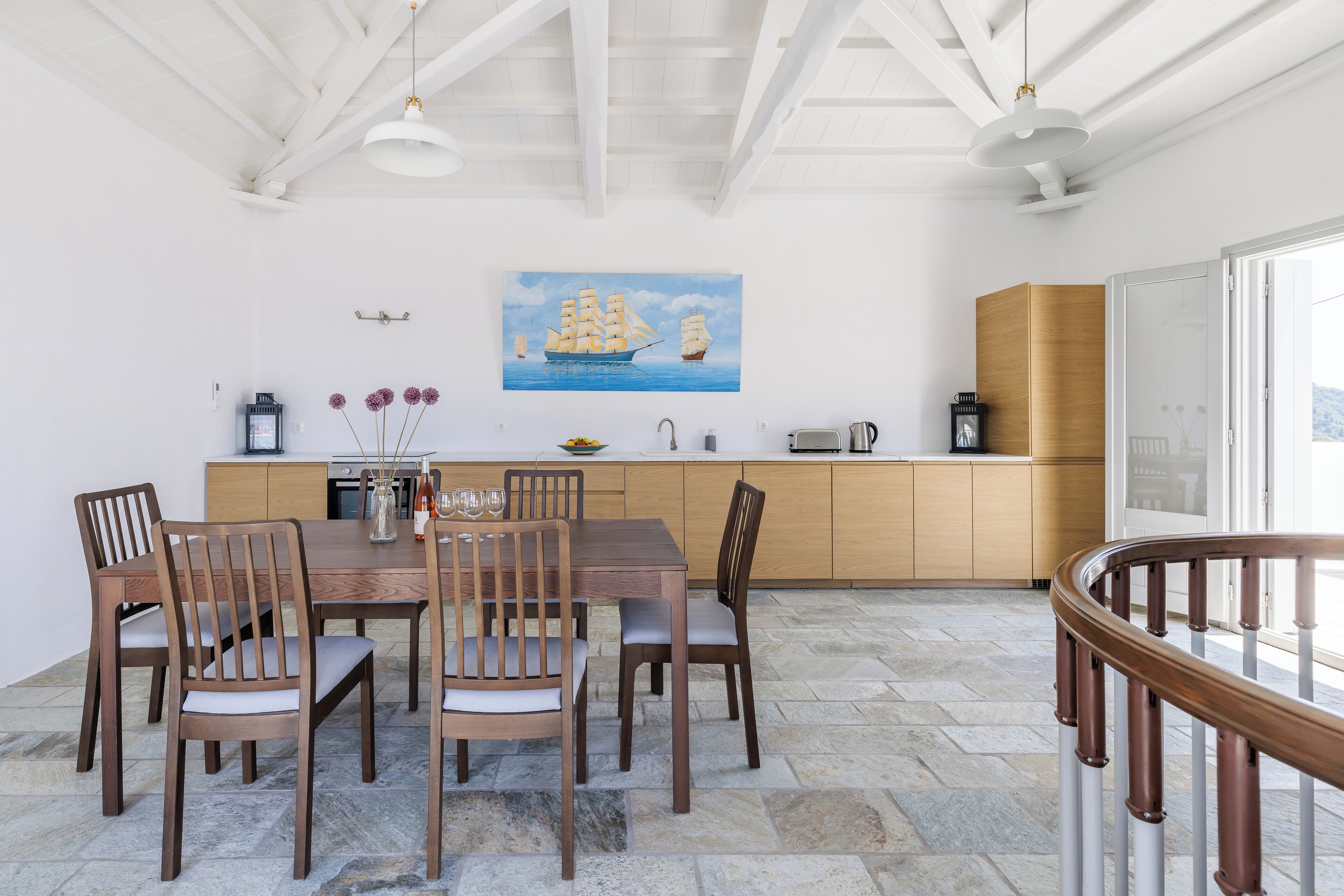 Un coin repas avec une table et des chaises en bois, un tableau représentant un navire accroché au mur, et une cuisine en arrière-plan.