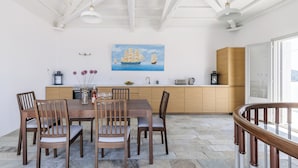 Uma área de refeições com uma mesa e cadeiras de madeira, um quadro de um navio na parede e uma cozinha ao fundo.