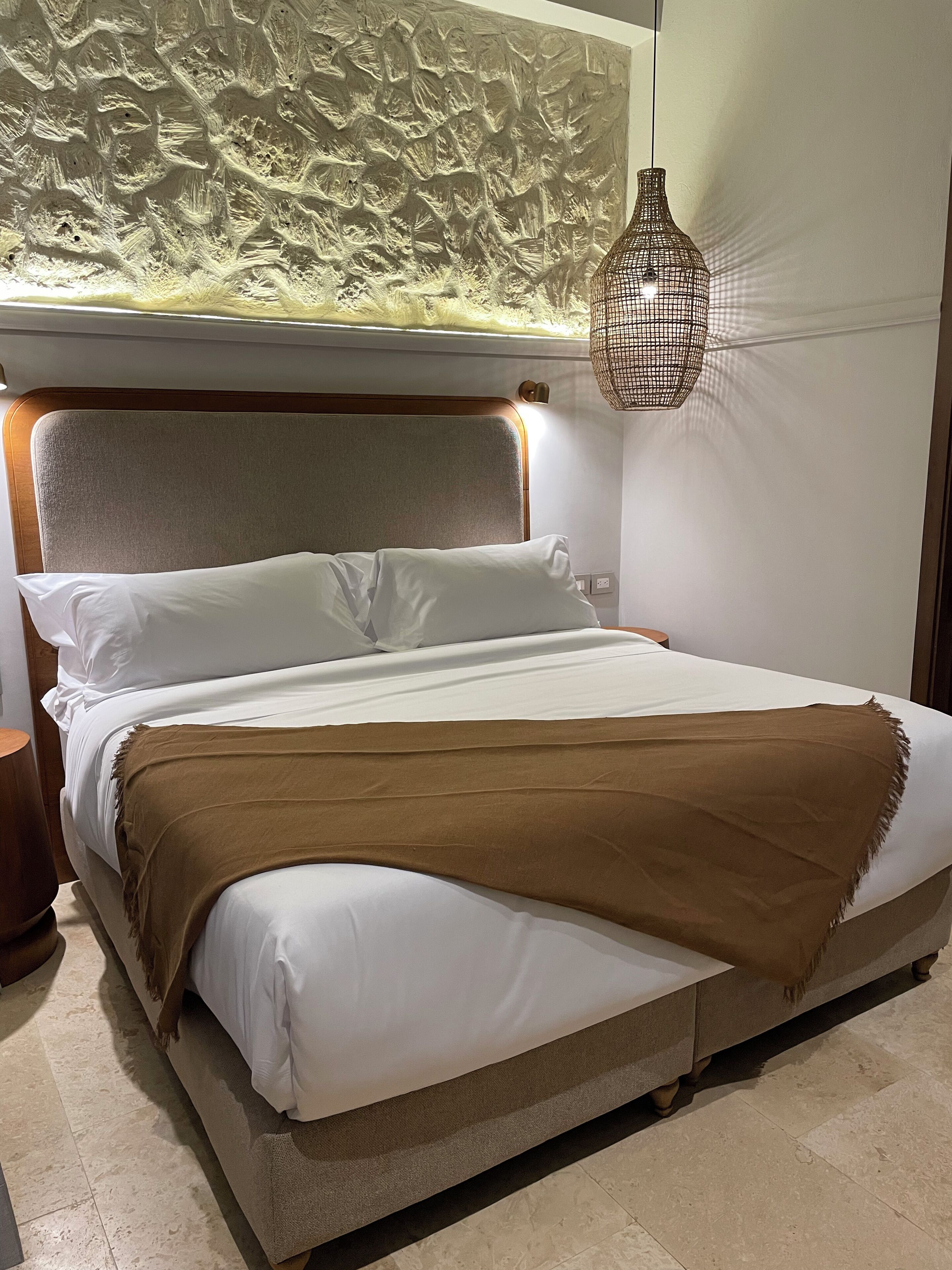 Ein Bett mit weißer und brauner Bettwäsche, eine strukturierte Wand dahinter und eine hängende Lampenleuchte.