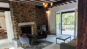 Un salon chaleureux avec une cheminée en pierre, des sièges confortables et une vue sur l’extérieur, accessible par une porte vitrée.