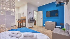 Une chambre d’hôtel moderne équipée d’un lit, d’un bureau, d’une chaise, d’une télévision et d’une salle de bain visible à travers une porte ouverte.