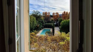Une vue à travers une fenêtre sur une piscine entourée d’arbres et d’arbustes.