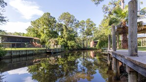 Een rustig waterlichaam met een houten steiger, omgeven door bomen en een gebouw met een overdekt terras.