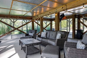 Un patio extérieur couvert, aménagé avec des meubles en osier, de grandes fenêtres et un plafond orné de poutres apparentes et illuminé de guirlandes lumineuses.