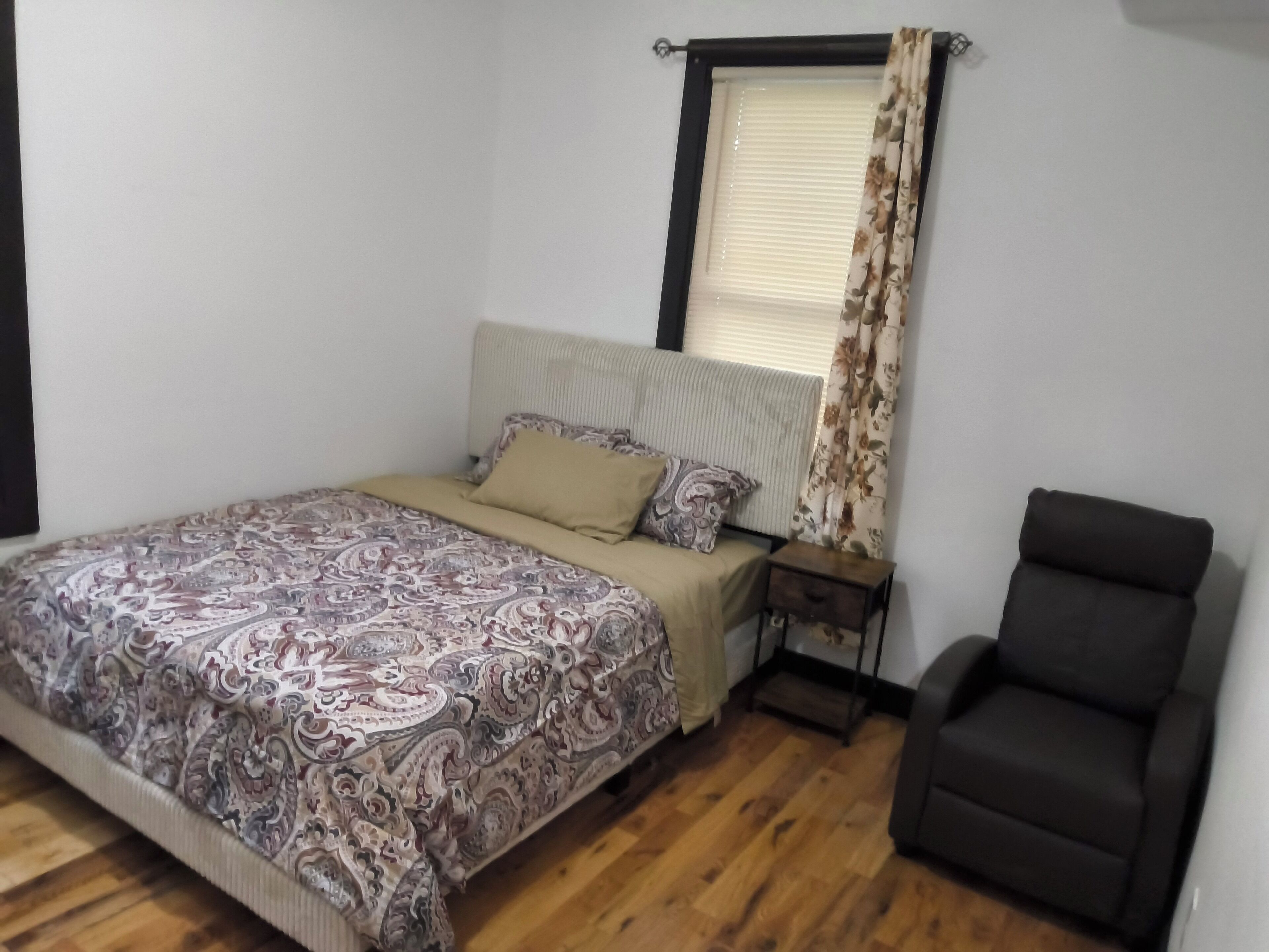 Un dormitorio con cama, mesita de noche, silla y ventana con cortinas.