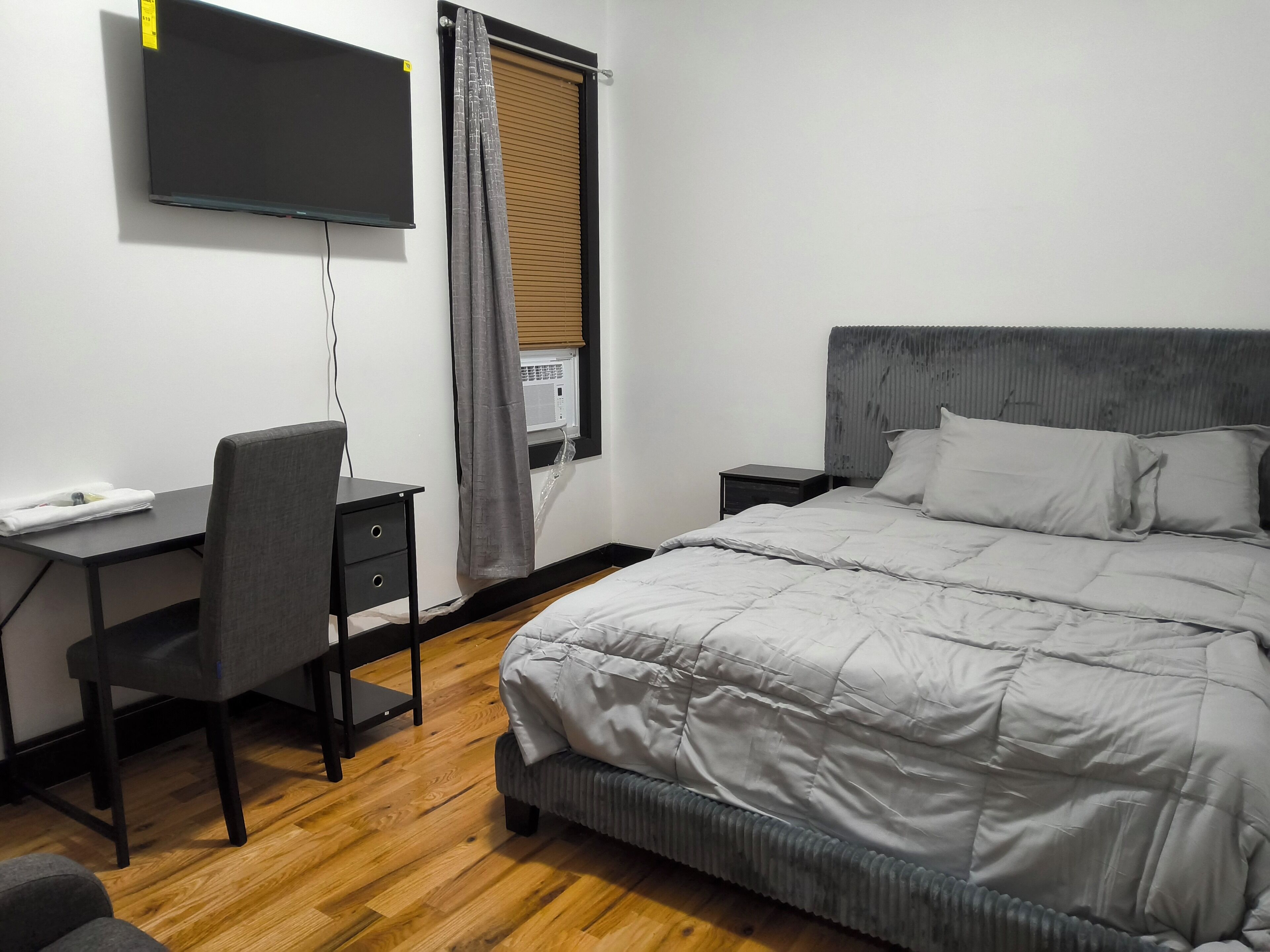 A bedroom with a bed, a desk, a chair, a TV mounted on the wall, and a window with blinds.