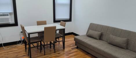 Une pièce avec un plancher en bois, une table blanche et quatre chaises, un canapé gris et deux fenêtres équipées de stores.