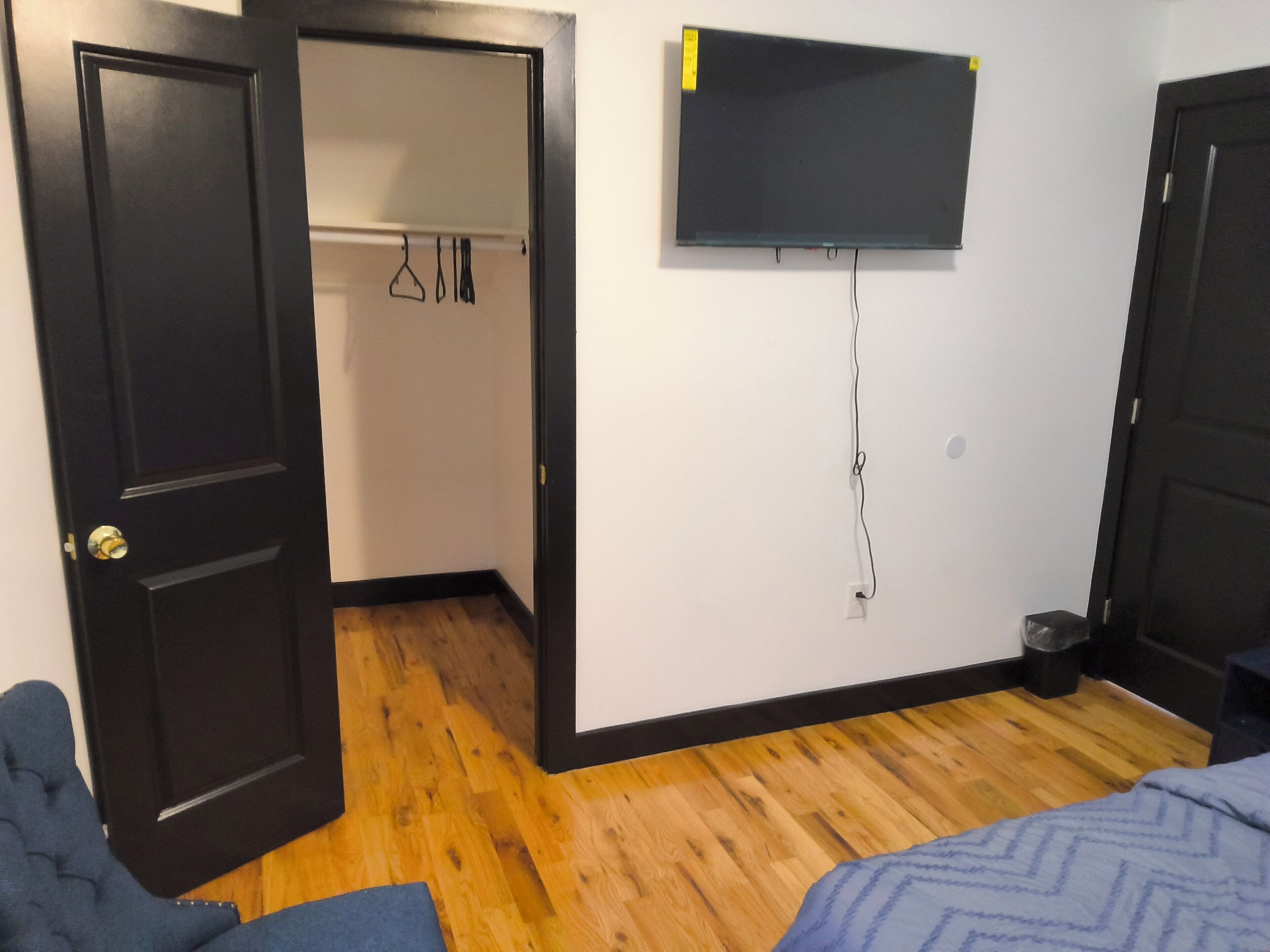 A room with a wooden floor, a bed, a TV mounted on the wall, and a door leading to another room.