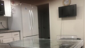 Eine moderne Küche mit Esstisch und Glasplatte, transparenten Stühlen, einem weißen Kühlschrank, einer Uhr und einem an der Wand befestigten Fernseher.
