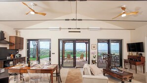 A spacious living area with a dining table, chairs, a sofa, and a TV. There are ceiling fans, a fireplace, and a view through large glass doors. - Private Hilltop Villa with Sea Views in the Heart of Maremma, Tuscany (Scansano)