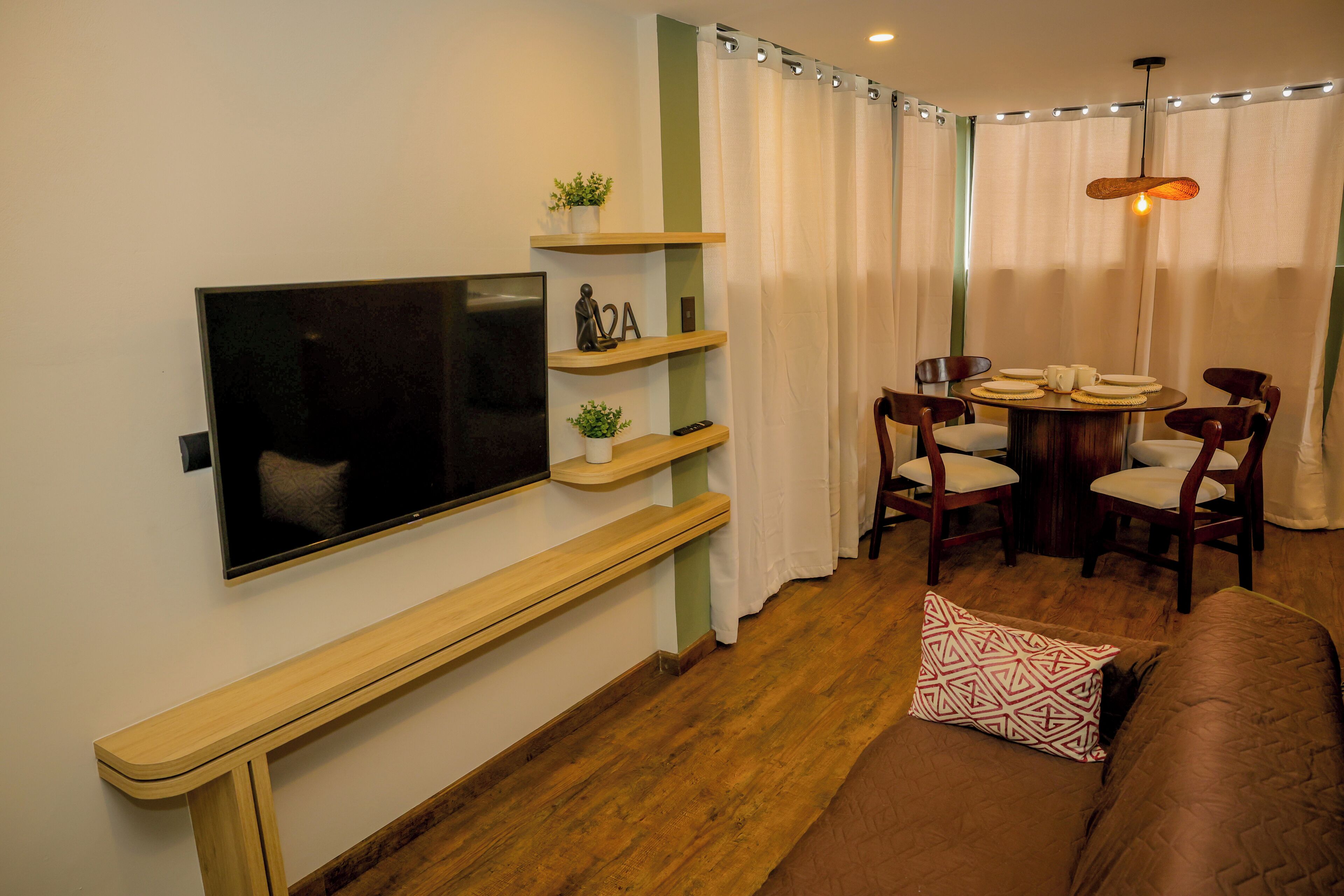 A modern living room with a flat-screen TV, wooden shelves, a round dining table with chairs, and a brown sofa.