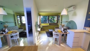 A modern living room with a sofa, coffee table, and dining area. There is a kitchen area with a counter and cabinets. The room has large windows and a view of the outdoors. - Cozy Apartment (Santa Tecla)
