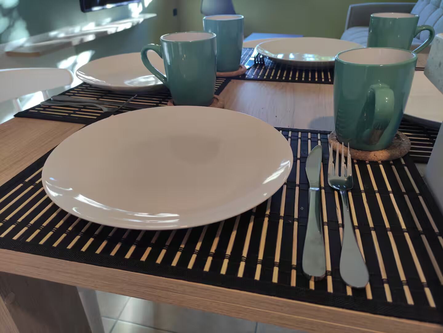 Una mesa de comedor dispuesta con un plato blanco, un tenedor y un cuchillo, junto a una taza verde.