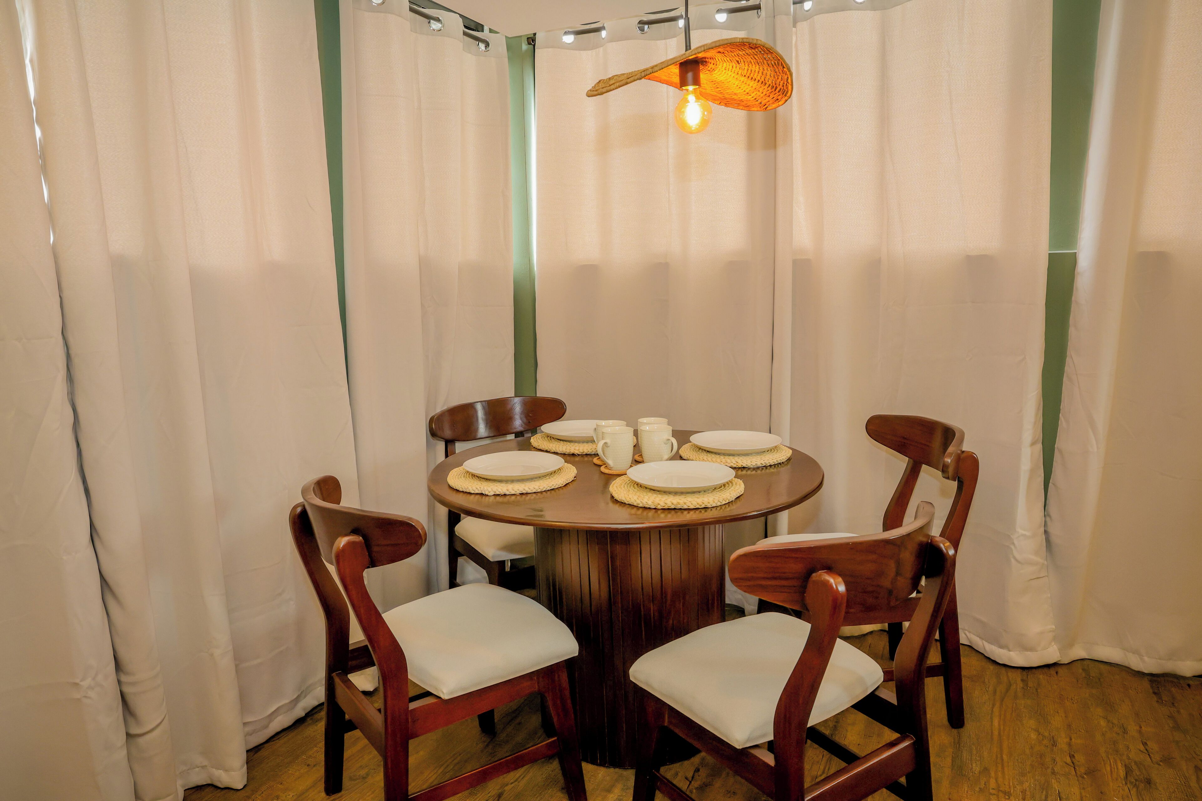 A round wooden dining table with white plates and cups, surrounded by four wooden chairs with white cushions, set against a backdrop of white curtains and a green stripe.