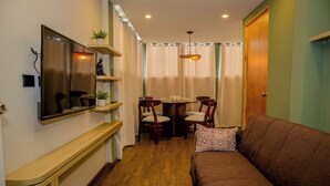 A compact living space with a sofa, a dining table with chairs, a TV mounted on the wall, and a wooden floor. - Cozy Apartment (Santa Tecla)