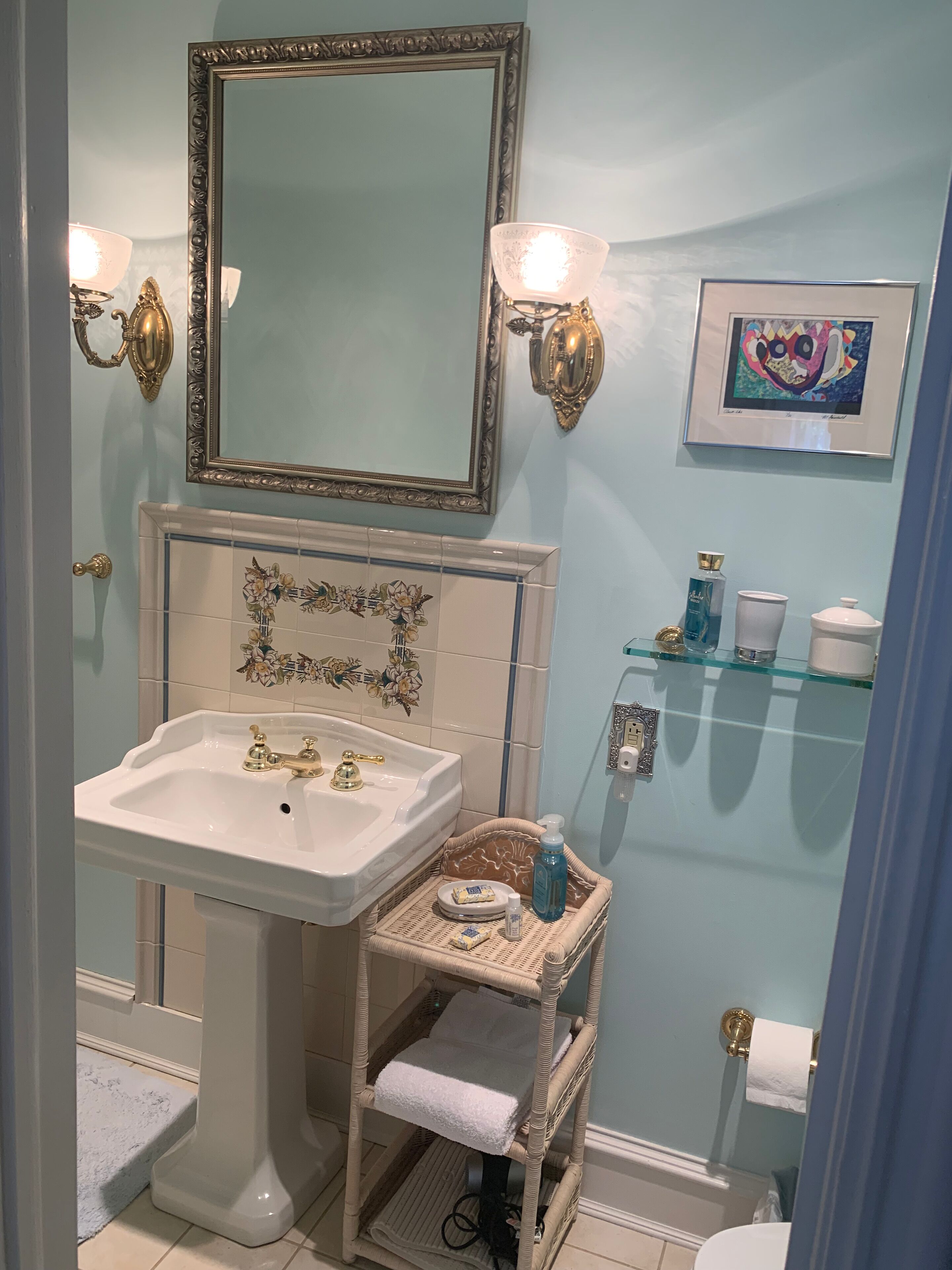 A bathroom with a pedestal sink, two wall-mounted lights, a mirror with an ornate frame, a wicker basket with toiletries, and a framed artwork on the wall.