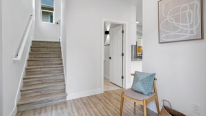 Un pasillo con una escalera de madera, una silla con cojín y una obra de arte abstracta enmarcada en la pared.