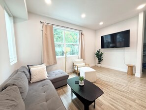 Un salon moderne avec un canapé d’angle, une table basse, un téléviseur fixé au mur et une fenêtre avec des rideaux.