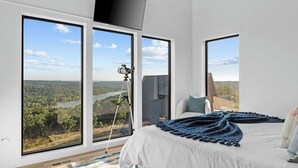 A bedroom with large windows, a bed, a mounted telescope, and a flat-screen TV. - Lux Lakeview • Pool, Hot Tub, Game Room, EpicViews (Omaha)
