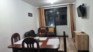 A dining area with a wooden table and chairs, a sofa, a TV mounted on the wall, and a window with curtains.
