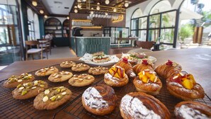 A variety of pastries and cookies on a table in a cafe with large windows and a brick wall in the background. - Tinspiro Alaçatı (Cesme)