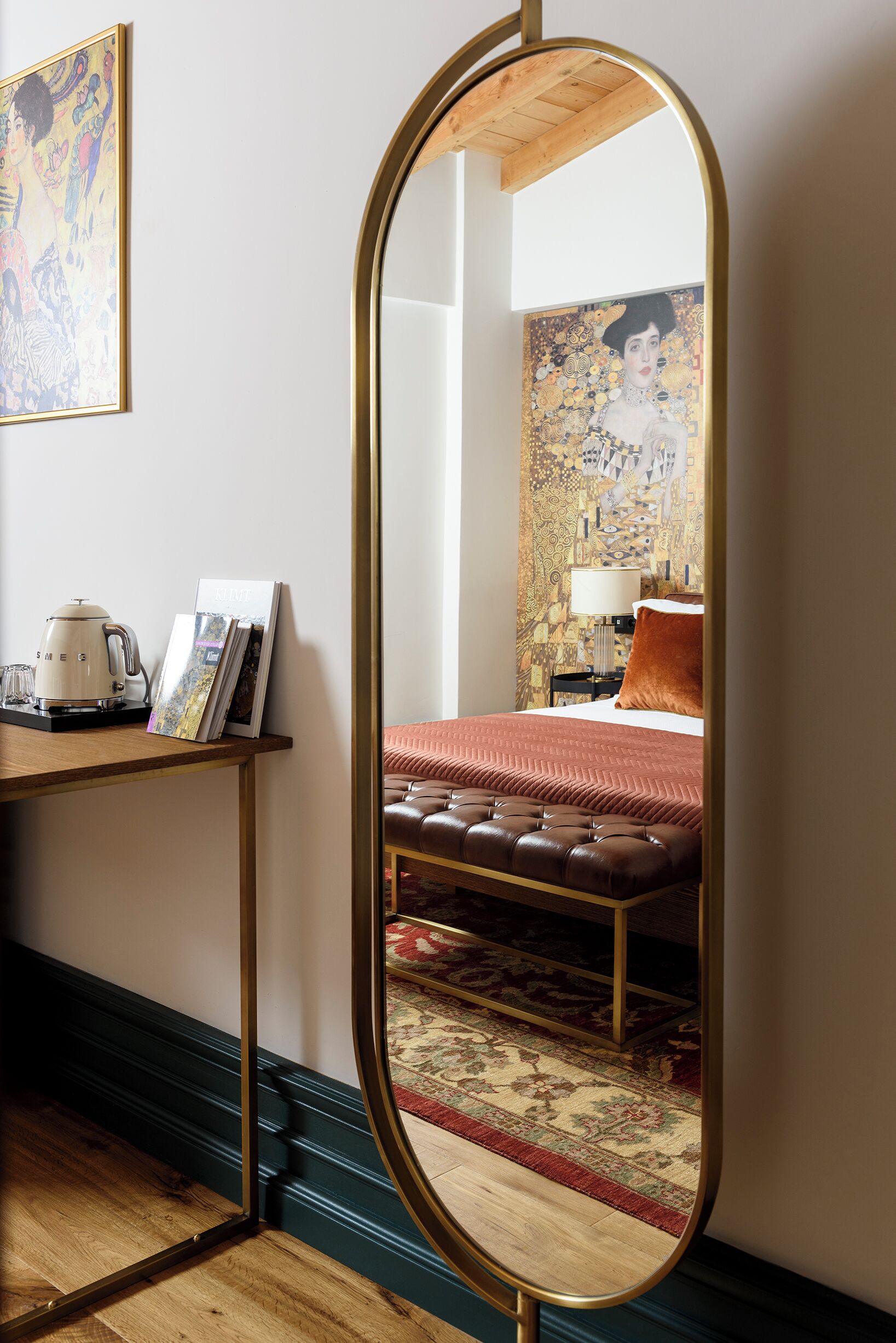 Habitació d'hotel amb un mirall gran, un llit amb col·cha vermella, una panxa de cuir, una cafetera i un quadre amb marc sobre una taula auxiliar.