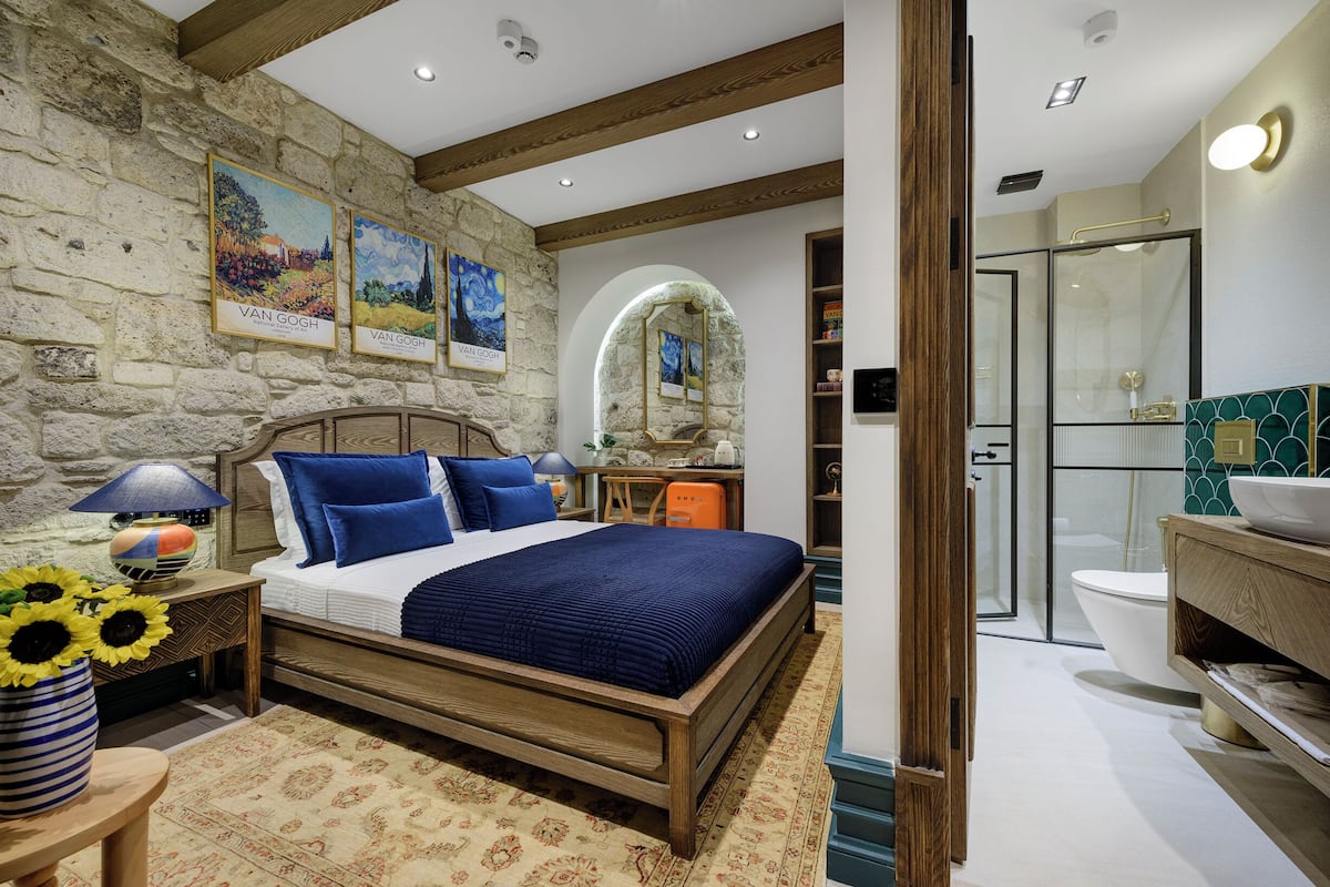 A bedroom with a stone wall, a wooden bed, a nightstand with a lamp, and a vase of sunflowers.