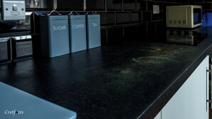 Eine Küche mit schwarzem Arbeitsplatz, drei beschriftete Behälter für Zucker, Kaffee und Tee, einer Mikrowelle und einer Steckdose.
