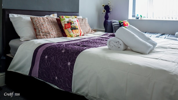 Ein ordentlich bezogenes Bett mit einer violett-weißen Tagesdecke, verschiedenen gemusterten Kissen und einem aufgerollten weißen Handtuch daneben.
