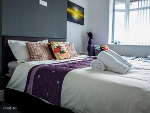 Uma cama bem arrumada com colcha roxa e branca, diversos travesseiros estampados e uma toalha branca enrolada ao lado.