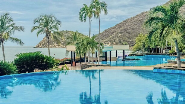A swimming pool with clear blue water, surrounded by palm trees and a view of the sea. - New KeyPöint: Long stays in Pözos Colorados! (Santa Marta)
