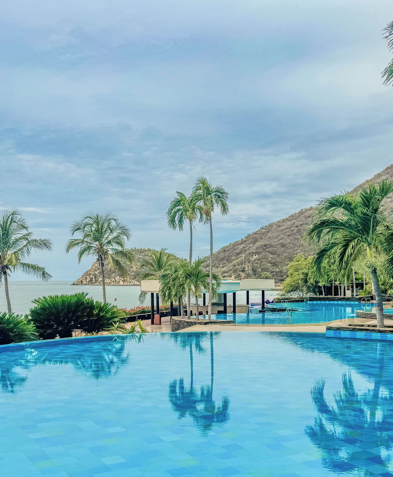 Ein Swimmingpool mit klarem, blauem Wasser, umgeben von Palmen und Blick auf das Meer.