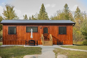 Una cabaña de madera con techo de metal, una escalera y un pequeño espacio exterior con mesa y sillas.