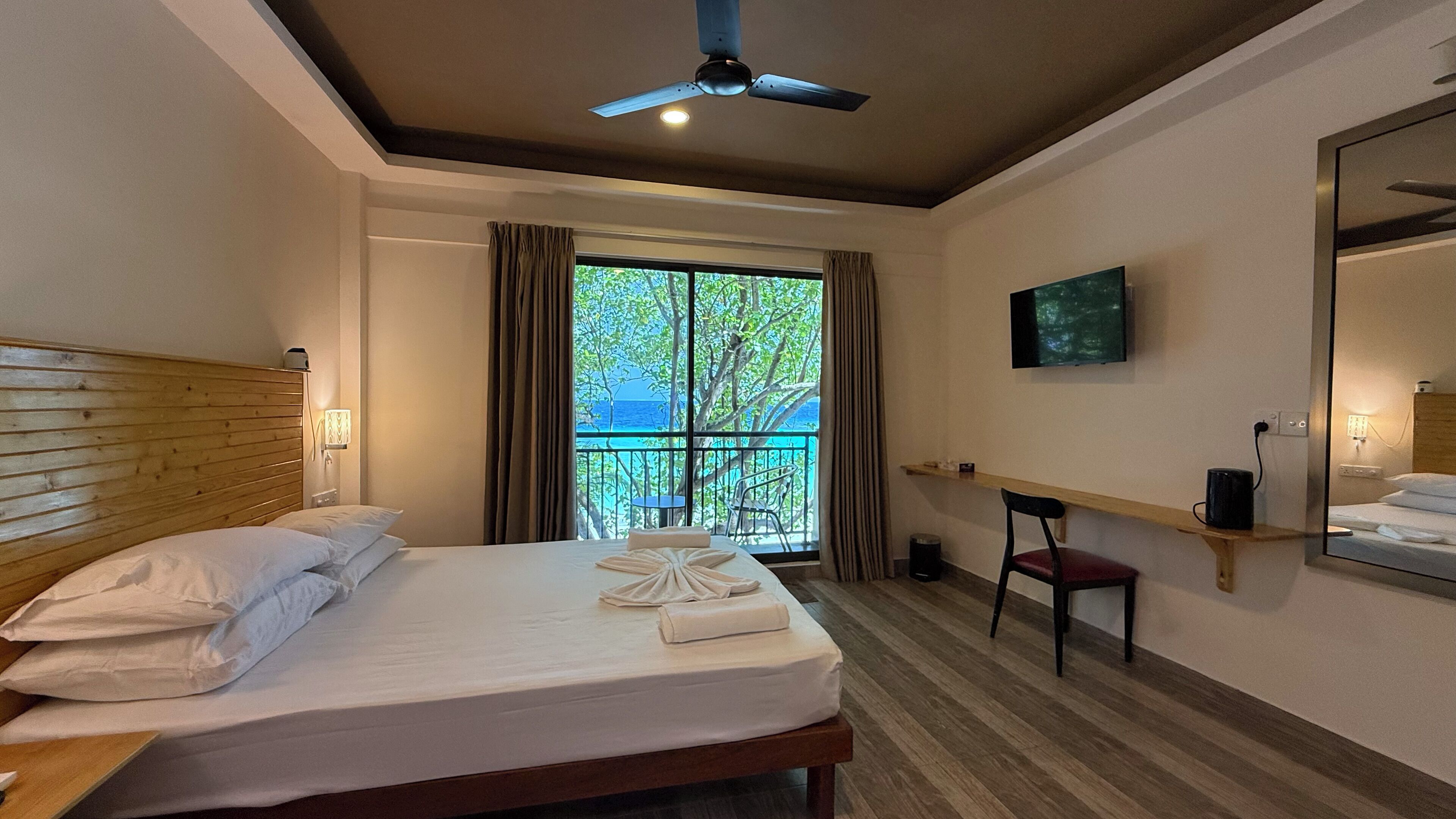 Una camera d'albergo con un letto, una scrivania, una sedia, una ventola a soffitto, una TV e un balcone con vista sul mare.