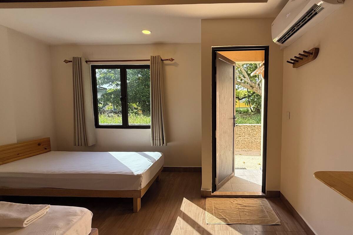 A bedroom with a wooden bed, a window with curtains, an open door to a balcony, and an air conditioning unit.