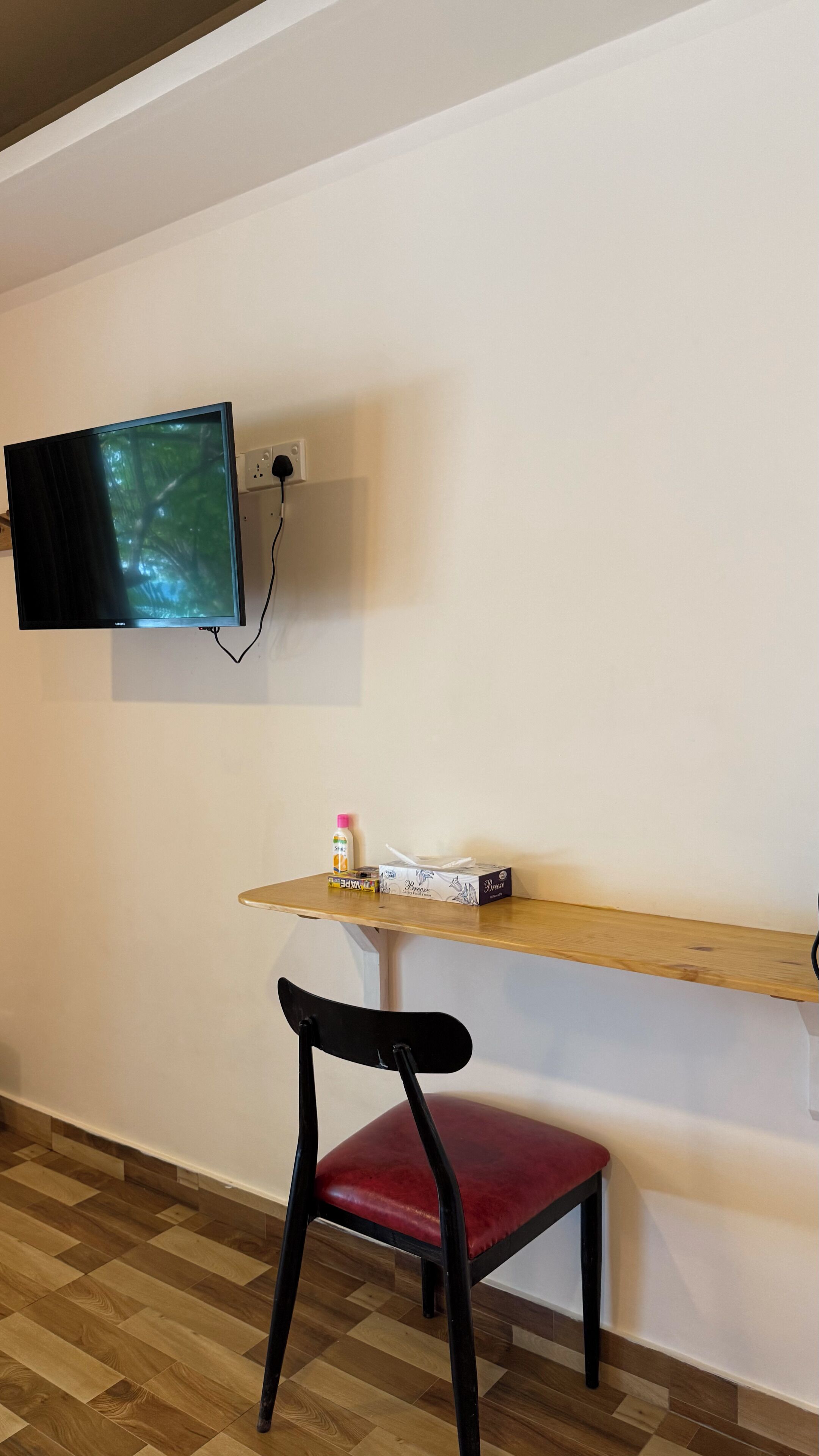 Ein Zimmer mit einem an der Wand befestigten Flachbildfernseher, einem Holzregal mit Taschentuchbox und einer Flasche sowie einem schwarzen Sessel mit rotem Kissen.