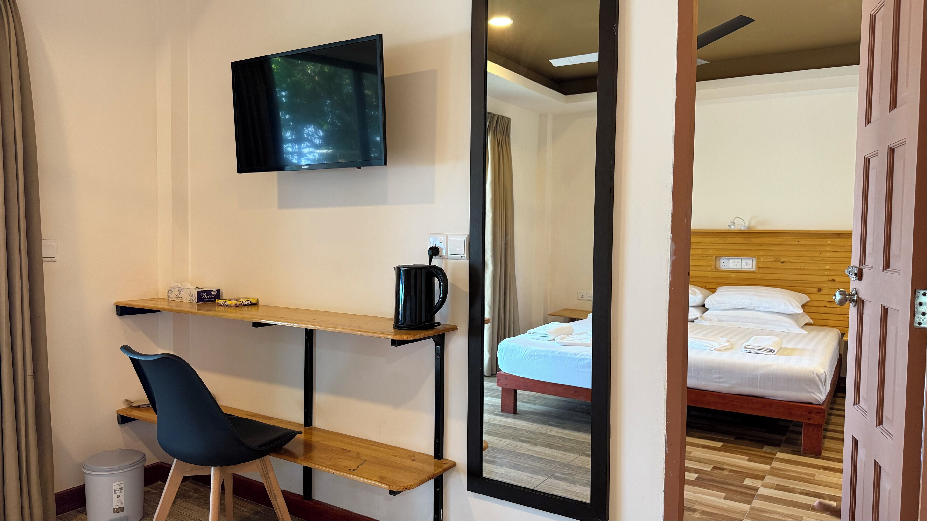 A hotel room with a bed, a desk with a chair, a TV, and a mirror reflecting the room.